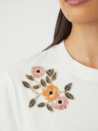 Hvit t-skjorte med blomsterbroderi av oransje og rosa blomster, sorte sentre og perleblad i mørk blå og gullakenter.
