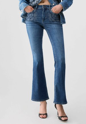 Donna che indossa jeans svasati blu con dettagli in strass sulle tasche, una giacca di jeans corta e sandali neri con tacco alto e punta aperta.