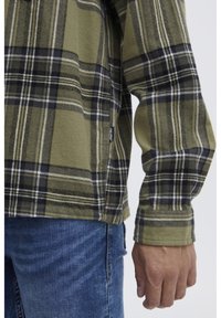 Grüne und schwarze karierte Flanellshirt mit langen Ärmeln, einer weichen Textur und einem Logosticker an der Seite, kombiniert mit blauen Jeans.