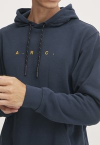 Sudadera con capucha de color azul marino con letras doradas "A.R.C.", cordón negro y puños acanalados. Textura suave y corte clásico.