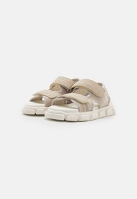 Beige sandaler med borrelås, nettingpaneler, en polstret såle og subtile sølvdetaljer. Rund tå og teksturert fotseng.