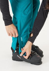 Turquoise skibroek met ritsopeningen, gecombineerd met zwarte skischoenen. Een lange mouw, donker shirt is zichtbaar, met duimgaten.