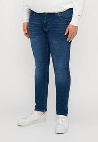 Jeans blu scuro a vestibilità slim, con classico design a cinque tasche, bottone in metallo e cuciture a contrasto; abbinati a sneakers bianche.