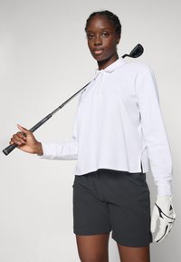 Vrouw in een witte langetermijn polo en zwarte shorts die een golfclub over haar schouder houdt en een witte golfhandschoen draagt op haar linkerhand.