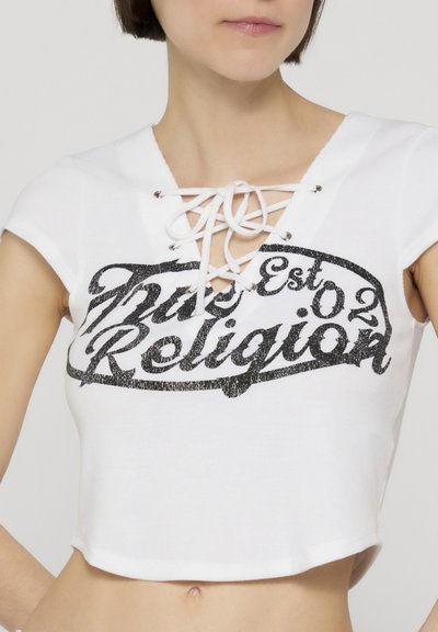Femme portant un top court blanc avec un col lacé et un texte noir "True Religion Est 02" sur la poitrine.