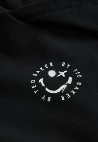 Sort stof med en blød tekstur, der har et rundt hvidt logo med et smilende ansigt, fly og sol, som visuelt er integreret i designet.