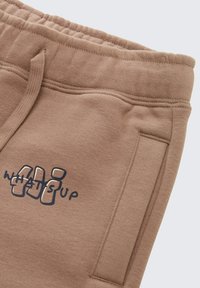 Lichtbruine katoenen joggingbroek met een elastische tailleband, zijzakken en een "WHAT'S UP" grafische opdruk in marineblauw. Soepele textuur.