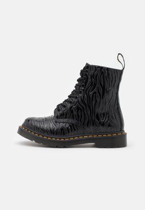 Botte de combat noire avec un motif ondulé texturé, huit œillets pour les lacets, coutures jaunes, et une languette à l'arrière, présentée sur un fond blanc.