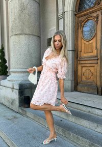 Femme en robe fleurie rose tenant une pochette blanche perlée, ajustant une sandale blanche à talon sur des marches en pierre devant un bâtiment avec porte en bois et colonnes.