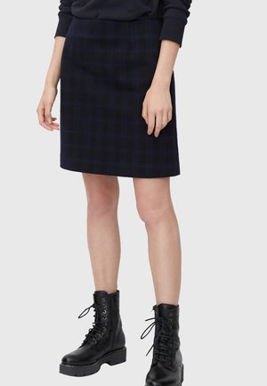 Femme portant une jupe écossaise foncée jusqu'aux genoux, des bottes de combat noires et un haut à manches longues sombre, debout devant un fond uni.