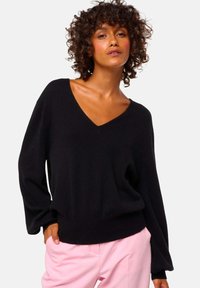 Pull en V noir avec des manches longues et décontractées, au toucher doux, associé à un pantalon rose clair, mettant en valeur un look décontracté.