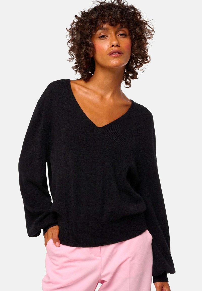 Pull en V noir avec des manches longues et décontractées, au toucher doux, associé à un pantalon rose clair, mettant en valeur un look décontracté.