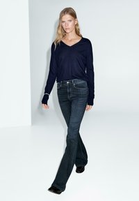 Marineblauwe langslijf V-hals top gecombineerd met donkerblauwe flare jeans. De stof lijkt zacht, en de outfit heeft een minimalistisch ontwerp en details.