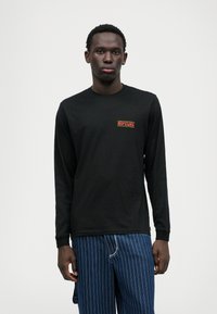 Czarna koszulka z długim rękawem z czerwoną naszywką "RIP CURL" na lewej piersi. Materiał jest gładki, a krój luźny.