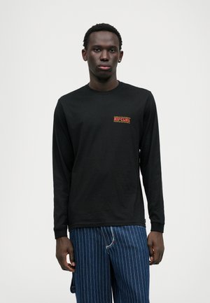 T-shirt noir à manches longues avec un logo rouge "RIP CURL" sur la poitrine gauche. Le tissu est lisse et la coupe est décontractée.