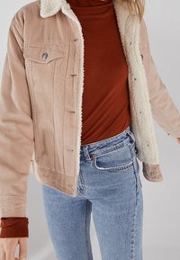 Giacca in corduroy con colletto in pelliccia sintetica color crema, chiusura anteriore con bottoni a pressione e due tasche sul petto, indossata sopra un dolcevita ruggine e jeans blu.