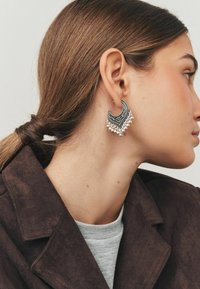 Metalgrå sølvøreringe med en halvmåneform, der har indviklede mønstre og hængende perleudsmykninger. Båret på en models øre mod en neutral baggrund.