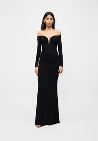 Γυναίκα που φοράει μακρύ μαύρο φόρεμα χωρίς ώμους με μακριά μανίκια και βαθύ V ντεκολτέ, στέκεται με τα χέρια χαλαρά στο πλάι.