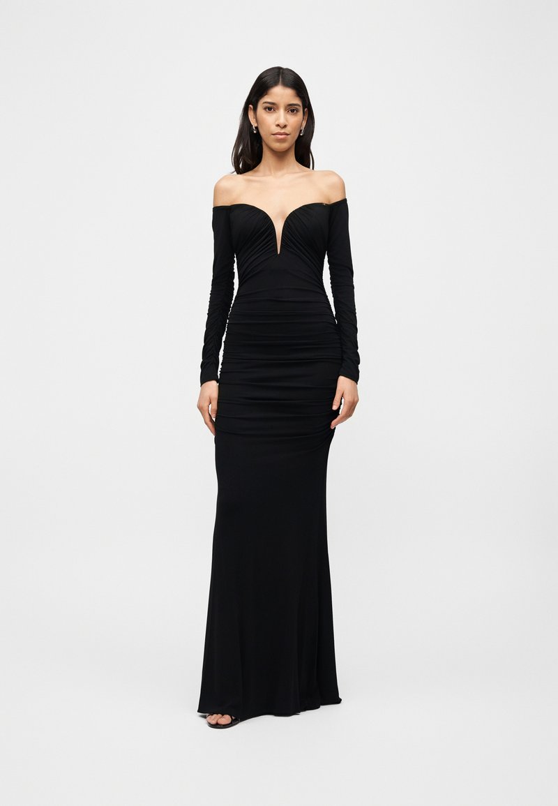 Γυναίκα που φοράει μακρύ μαύρο φόρεμα χωρίς ώμους με μακριά μανίκια και βαθύ V ντεκολτέ, στέκεται με τα χέρια χαλαρά στο πλάι.