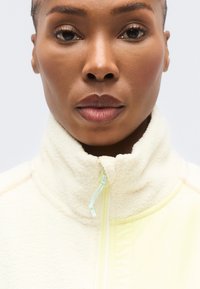 Ζακέτα από φλίς σε εκρού χρώμα με ψηλό γιακά, διαθέτει κλείσιμο με φερμουάρ, ανοιχτοπράσινες λεπτομέρειες και απαλή υφή, κατάλληλη για καθημερινή χρήση.