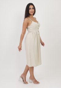 Beiges ärmelloses Kleid mit tailliertem Oberteil, Knopfdetails vorne und einem fließenden Rock; kombiniert mit klaren Blockabsatzsandalen.