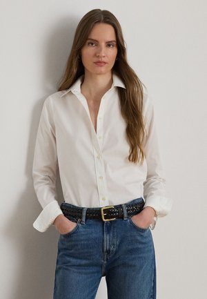 Žena s dlhými hnedými vlasmi, ktorá nosí bielu košeľu na gombíky zasunutú do modrých džínsov s čiernym pleteným opaskom, ruky v vreckách, neutrálna mimika.