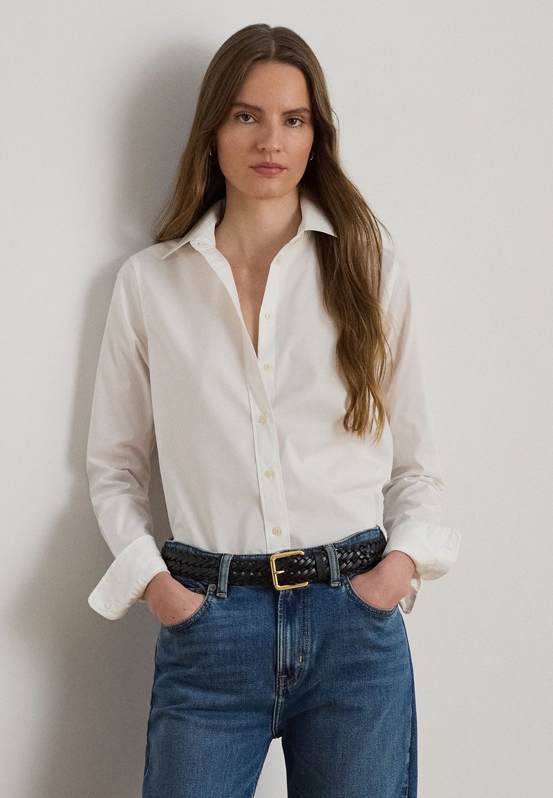 Žena s dlhými hnedými vlasmi, ktorá nosí bielu košeľu na gombíky zasunutú do modrých džínsov s čiernym pleteným opaskom, ruky v vreckách, neutrálna mimika.