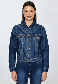 Moteris su tiesiais rudos spalvos plaukais, dėvi auksinius žiedinius auskarus, segėtą tamsiai mėlynos džinsinio audinio striukę ir derančius tamsiai mėlynos džinsinio audinio džinsus.