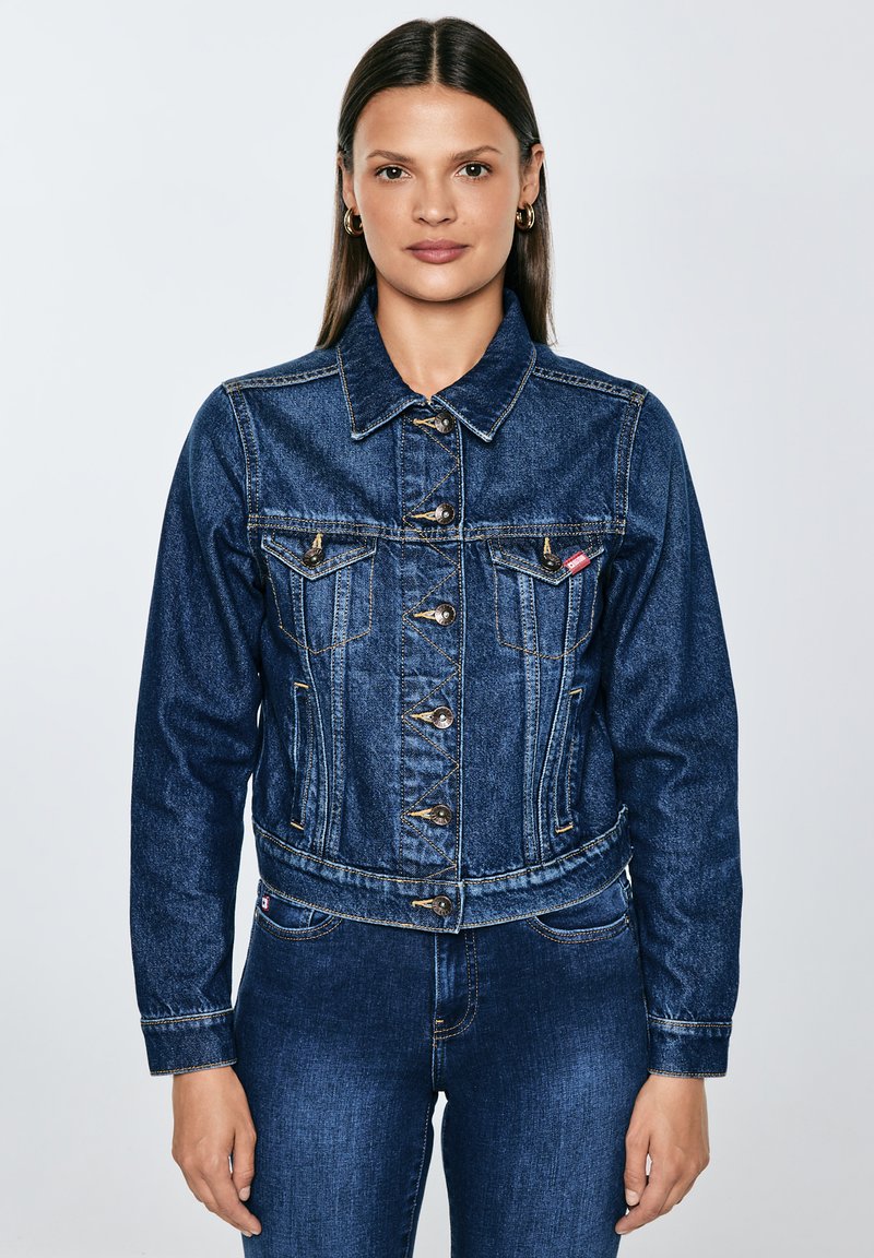 Moteris su tiesiais rudos spalvos plaukais, dėvi auksinius žiedinius auskarus, segėtą tamsiai mėlynos džinsinio audinio striukę ir derančius tamsiai mėlynos džinsinio audinio džinsus.