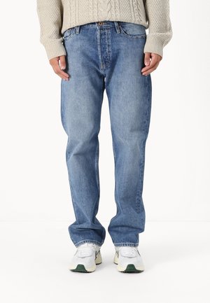 Person trägt locker sitzende hellblaue Jeans aus Denim, einen cremefarbenen Pullover mit Zopfmuster und weiße Turnschuhe mit grünen Akzenten, steht auf weißem Boden.