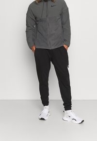 Giacca con cappuccio Nike grigia, chiusura con zip, pantaloni jogger neri con logo bianco, scarpe da ginnastica bianche con accenti neri, vestibilità casual.