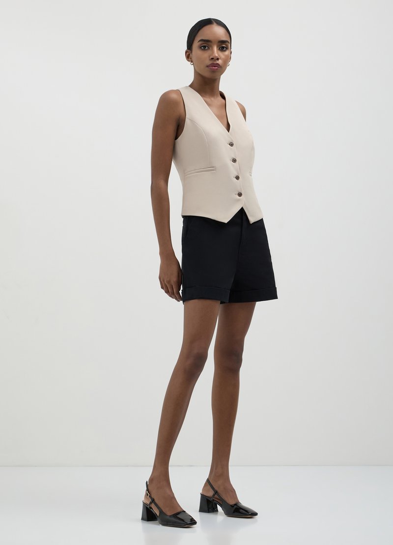 Gilet sans manches beige avec un col en V, cinq boutons sur le devant et des poches latérales, associé à un short noir et des sandales à talons noirs.
