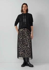 Femme portant un top à manches longues noir avec des liens en ruban, une jupe plissée à imprimé léopard et des bottines noires à la cheville, se tenant contre un mur uni.