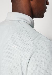 Hellblaues Poloshirt mit einem dezenten Allover-Muster, geknöpftem Kragen und einem kleinen weißen Logo auf der Rückseite in der Nähe des Halsausschnitts.