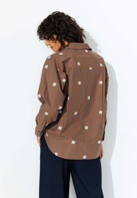 Chemise à rayures brunes avec broderie florale blanche. Manches longues, col, coupe loose. Portée avec un pantalon à rayures bleu marine.