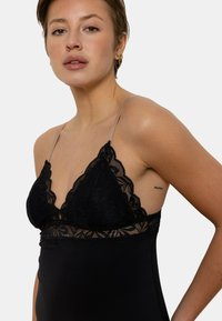 Bralette en dentelle noire avec des bonnets triangulaires, des bretelles ajustables transparentes, un motif floral et un tissu lisse. Présente un design ajusté.