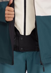 Heren zip-up jack in teal en wit met een zwarte binnenkant. Voorzien van een staande kraag, elastische manchetten en drukknopen aan de binnenkant. Vervaardigd uit glad, waterafstotend materiaal.