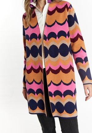 Long cardigan avec un motif coquille géométrique audacieux en rose, orange, marine et bordeaux sur une chemise blanche et un pantalon noir.