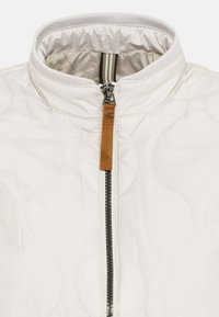 Weiß gesteppte Jacke mit hohem Kragen, ausgestattet mit einem Vorderreißverschluss mit einem braunen Lederzipper und kontrastierendem gestreiftem Stoff im Inneren des Kragens.