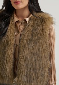 Femme portant un gilet en fausse fourrure marron sur une chemise plissée, brillante, marron clair, boutonnée, avec des cheveux châtain foncé visibles.