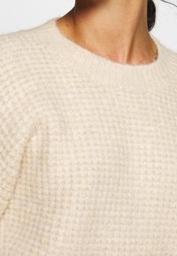 Pull en maille beige clair avec un motif gaufré texturé et un col rond côtelé, offrant une apparence douce et confortable.