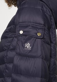 Granatowa kurtka puchowa z pikowaną teksturą, złotymi akcentami i naszywką z logo "RLL". Posiada zamykaną na zamek kieszeń na ramieniu.