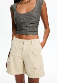 Grå spetscroptopp med rund halsringning, tillsammans med beige cargo-shorts som har sidofickor och metalldetaljer. Tyget ser mjukt och texturerat ut.