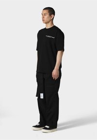 Svart t-skjorte med kortermet og hvit logo på brystet, kombinert med løse svarte cargobukser med lommer og etiketter. Svart sneakers med hvite snøring.