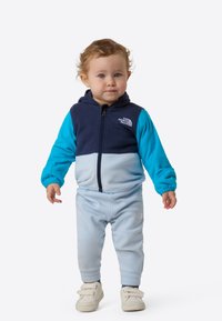 Jachetă din fleece cu fermoar, albastru deschis și bleumarin, cu glugă, mâneci lungi și design bicolor. Asortată cu pantaloni albastru deschis.