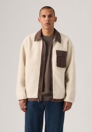 Cream fleece jacket with dark brown collar and chest pocket; features zip closure and textured surface; paired with a gray shirt and denim.