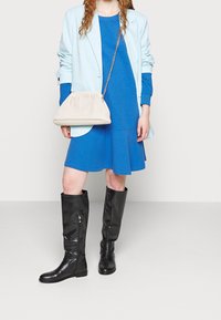 Vaaleansininen blazer sinisen mekon päällä, yhdistettynä mustiin polvimittaisiin tekstuuribootseihin ja väriltään kermaiseen käsilaukkuun, jossa on neulottu muotoilu ja ketjuhihna.