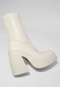 Valkoinen nahkainen nilkkurit korkean, paksun korokkeen kanssa, pyöristetty kärki ja tyylikäs, sileä pinta. Sisältää vähäistä ompelua sauman varrella.