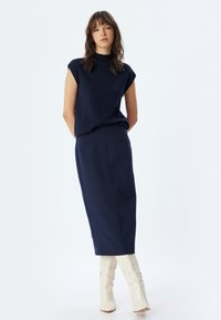 Marineblaue ärmellose Bluse, kombiniert mit einem knielangen marineblauen Rock, beide aus glattem Stoff. Das Model trägt weiße High-Heels und steht vor einem hellen Hintergrund.