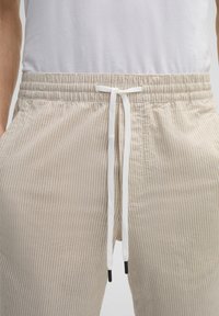 Beige gestreifte Baumwollshorts mit einem elastischen Bund, weißem Kordelzug und Seitentaschen. Der Stoff hat eine strukturierte Oberfläche.